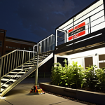 **

A rooftop garden scene. Focus on a clearly marked, well-lit emergency escape route (stairs or ladder) leading down from the roof. Include visible safety signage (glowing in the dark). Ensure weather-resistant materials (steel, composite) are used for the escape route. A family doing an evacuation drill.
**